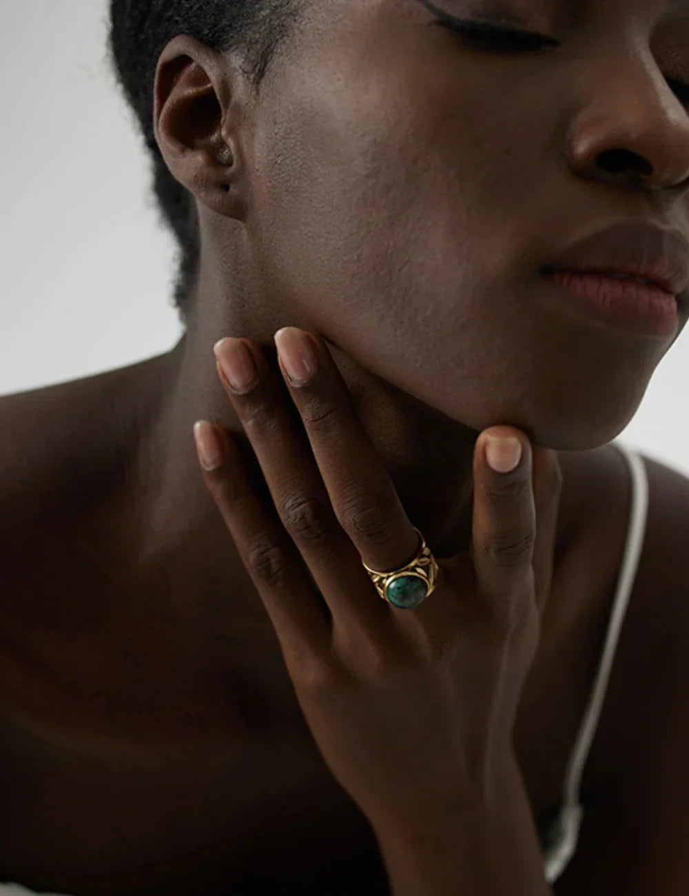 A close-up of a woman with dark skin and closed eyes, gently touching her neck while wearing the Turquoise Statement Ring—a gold plated piece with a large green stone—styled elegantly with a white thin-strap top for a bohemian look.