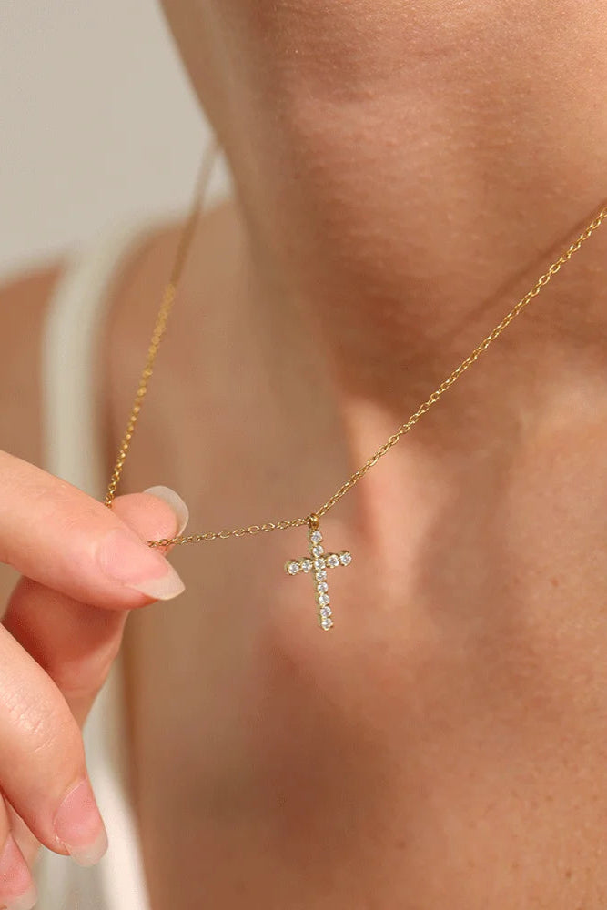 A close-up of a person gently lifting the Grace Cross Pendant Necklace with sparkling stones near their neck. The gold plated necklace shines against their white top.