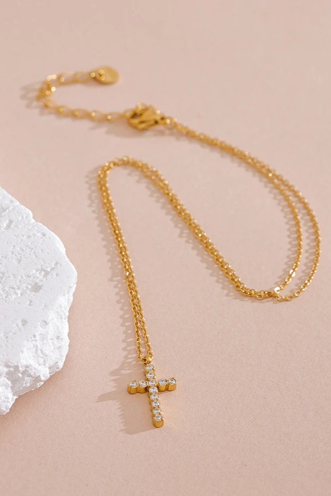 The Grace Cross Pendant Necklace, featuring a gold-plated cross accented with clear cubic zirconia stones, is shown on a light beige surface beside a textured white stone.