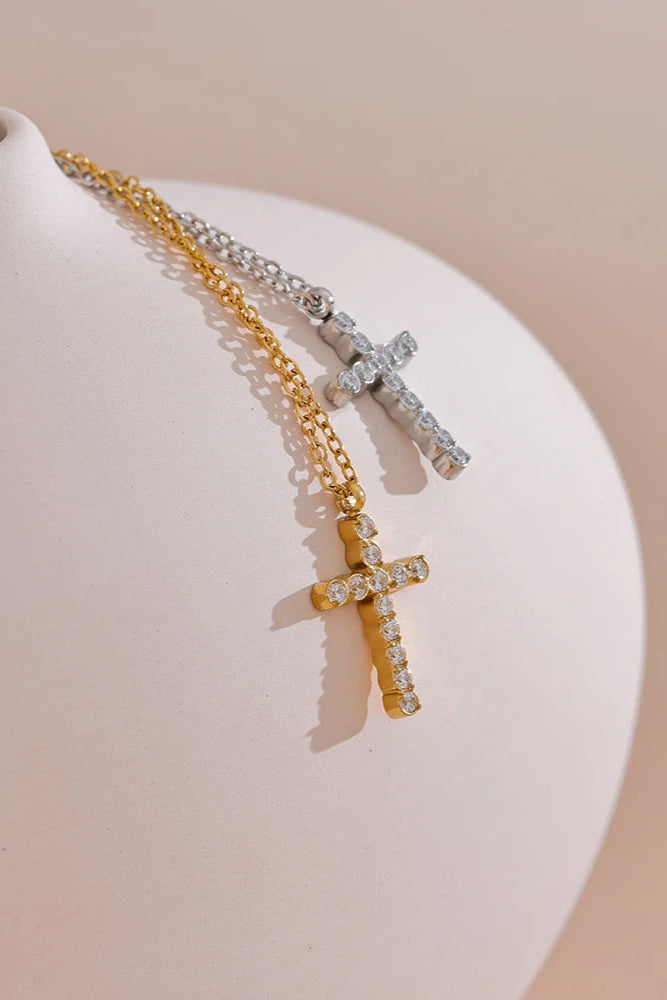 Two Grace Cross Pendant Necklaces, one gold plated and one silver, both with sparkling cubic zirconia, are displayed on a smooth light-colored curved surface against a soft beige background.